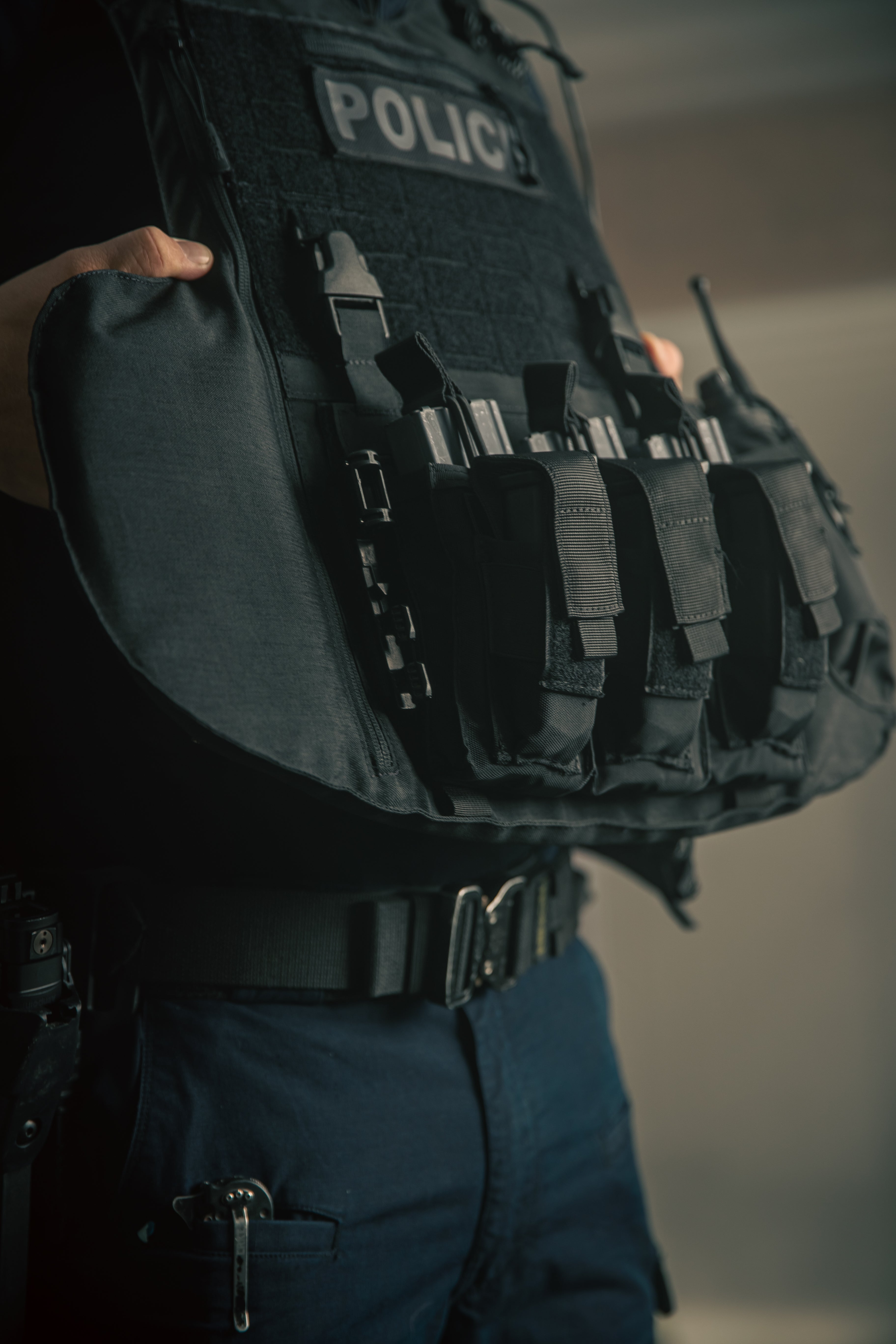 Close-up of a person wearing a dark tactical vest labeled Police, holding the sides. The vest has multiple pockets, conveying a sense of readiness.