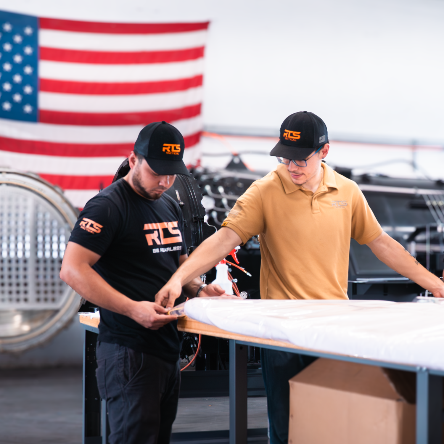 Two men in branded caps and shirts work intently in a workshop. An American flag hangs in the background, creating a focused, patriotic setting.