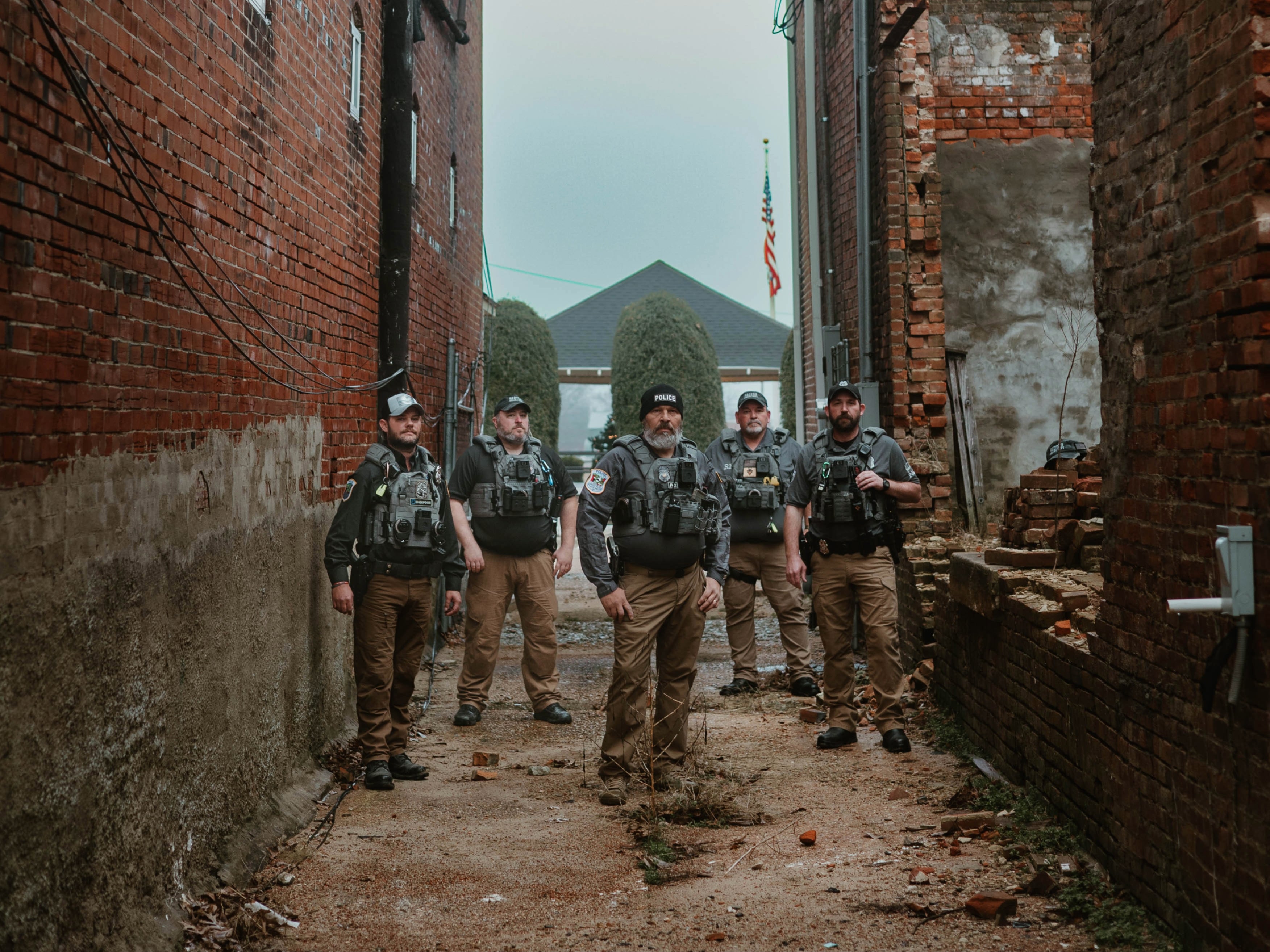 A group of six uniformed officers stands in a narrow alley between brick buildings. The mood is serious. An American flag is visible in the background.