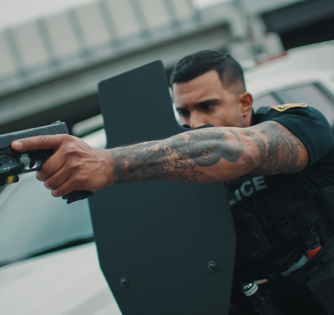 A police officer with a tattooed arm aims a handgun behind a ballistic shield, evoking tension and focus. A blurred police vehicle is in the background.