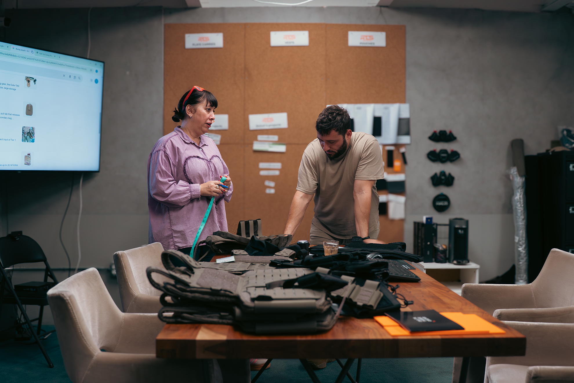 Two people stand in a workshop with tactical gear spread on a table. A screen displays items, and a bulletin board is visible. Focused and collaborative mood.