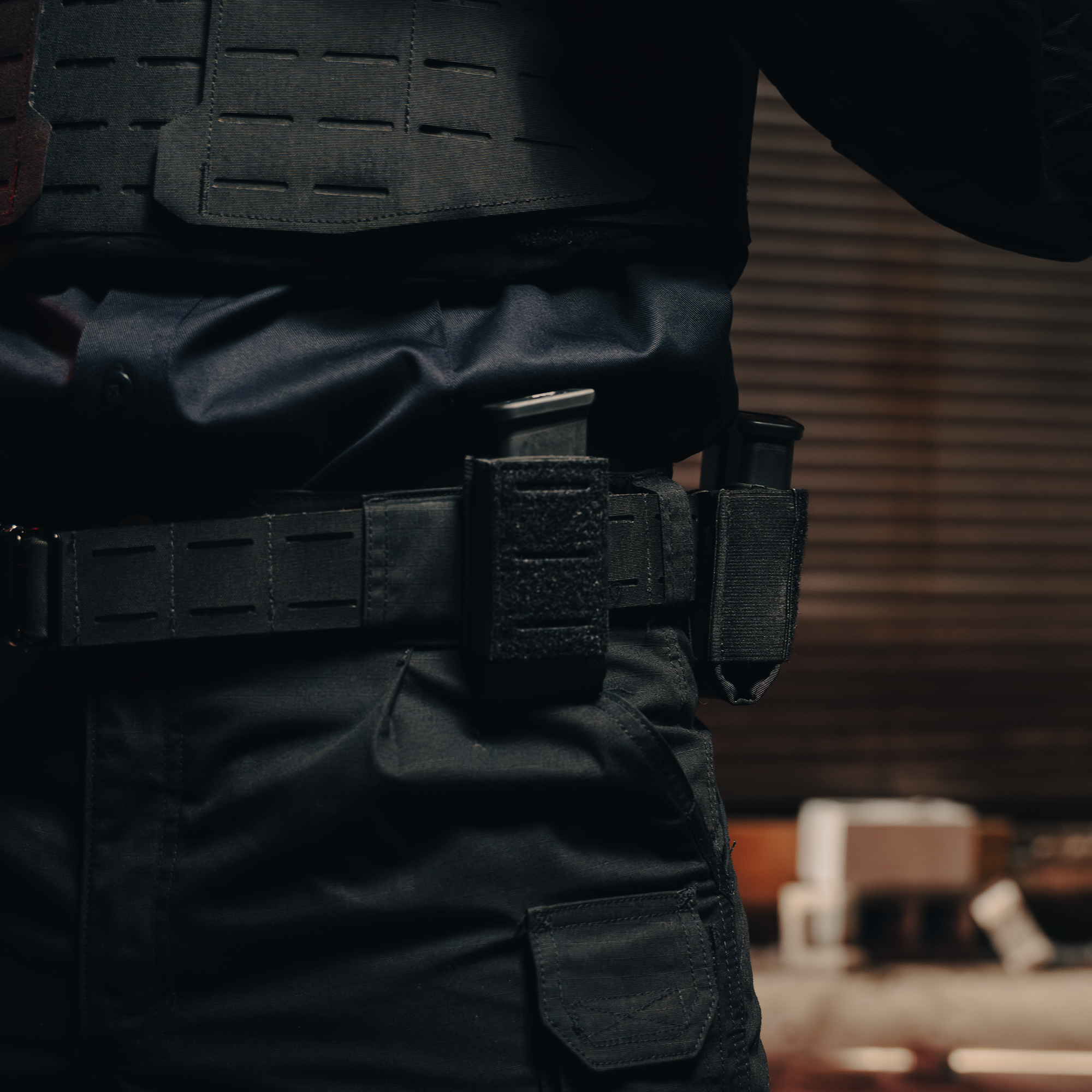 Close-up of a person wearing a dark tactical vest and pants, with a black utility belt holding gear. The background is blurred, suggesting an indoor setting.