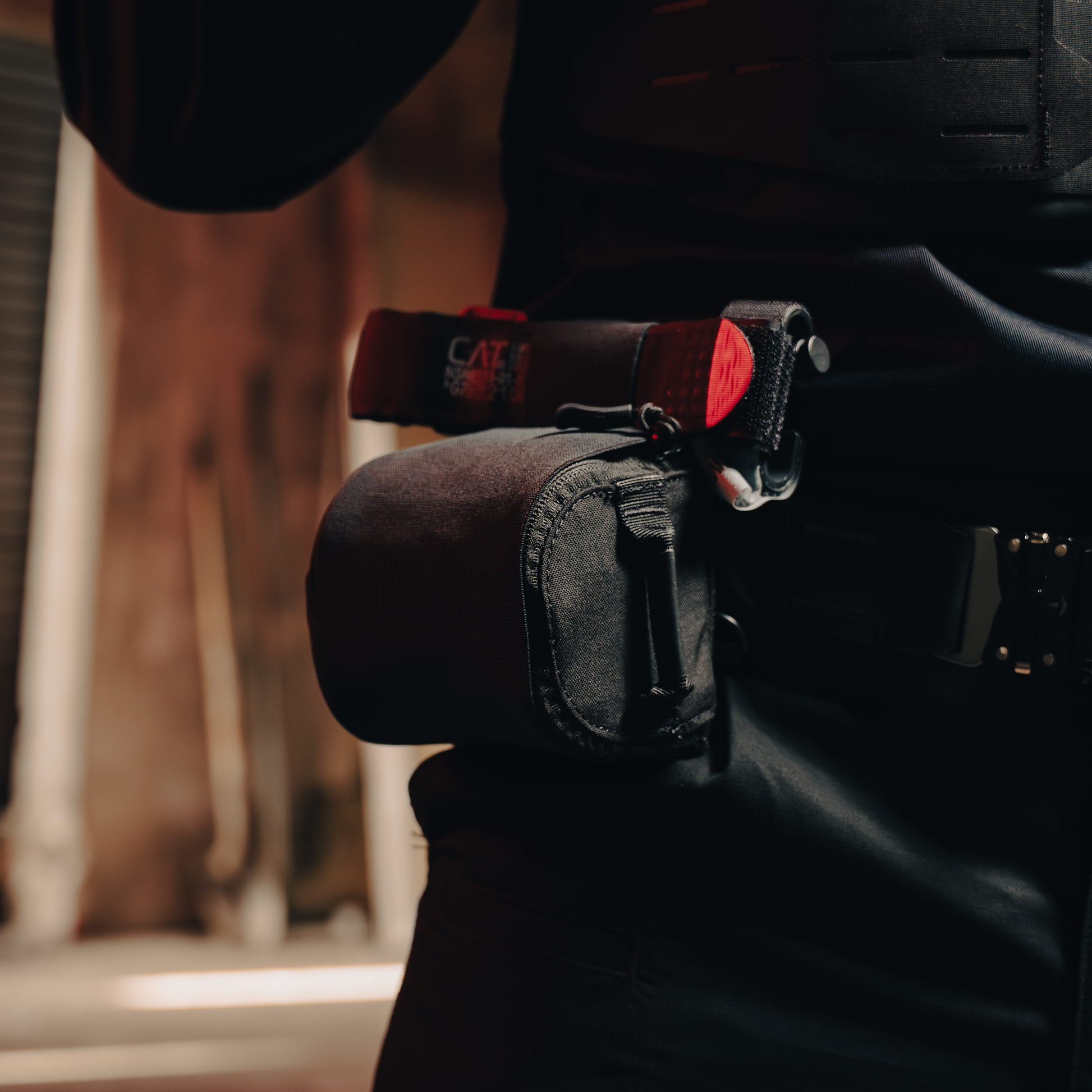 A close-up of a person wearing tactical gear, including a black equipment pouch and a red and black tourniquet on their belt, in a dimly lit room.