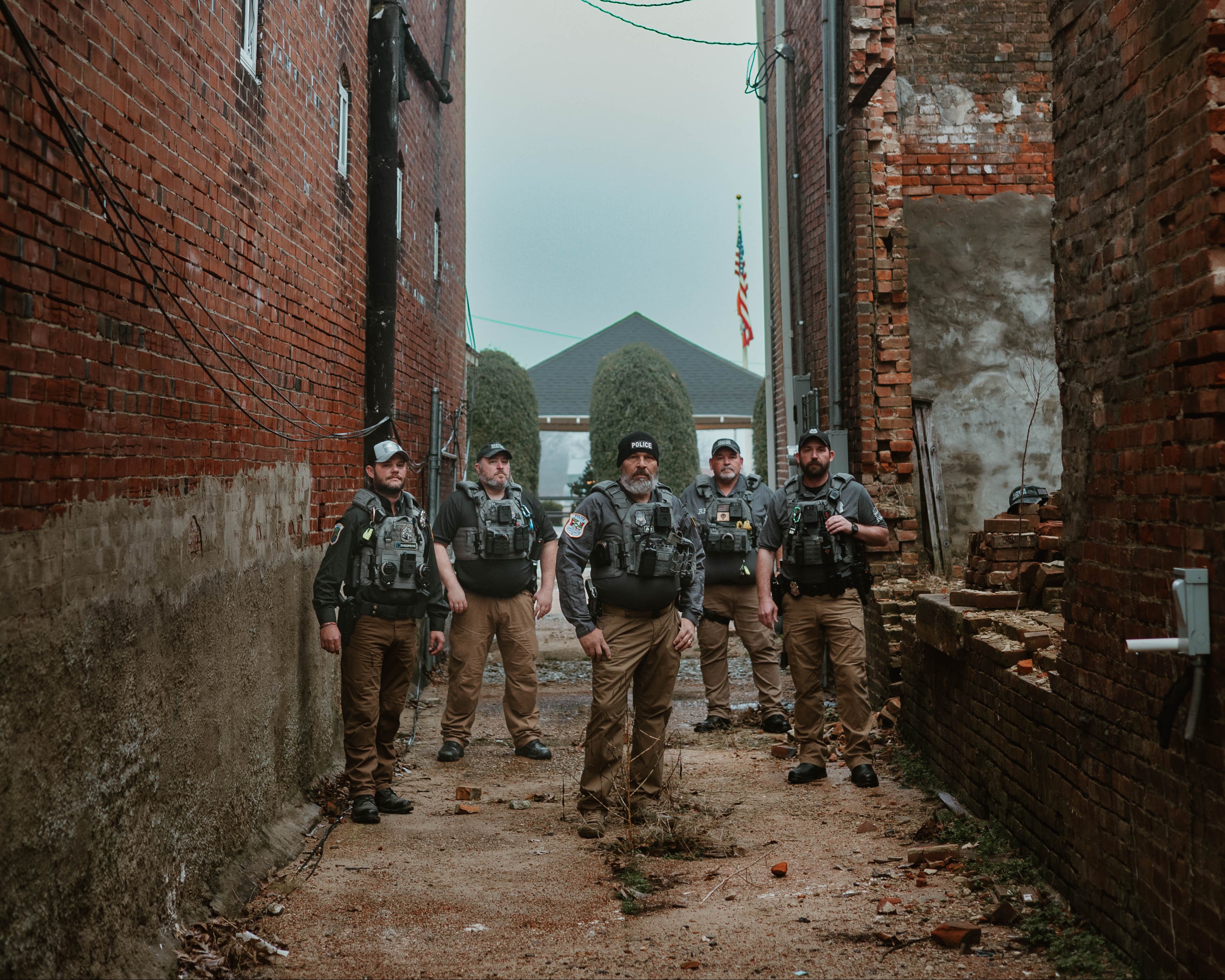 Five police officers in RTS tactical gear are standing in a narrow alley between two brick buildings.