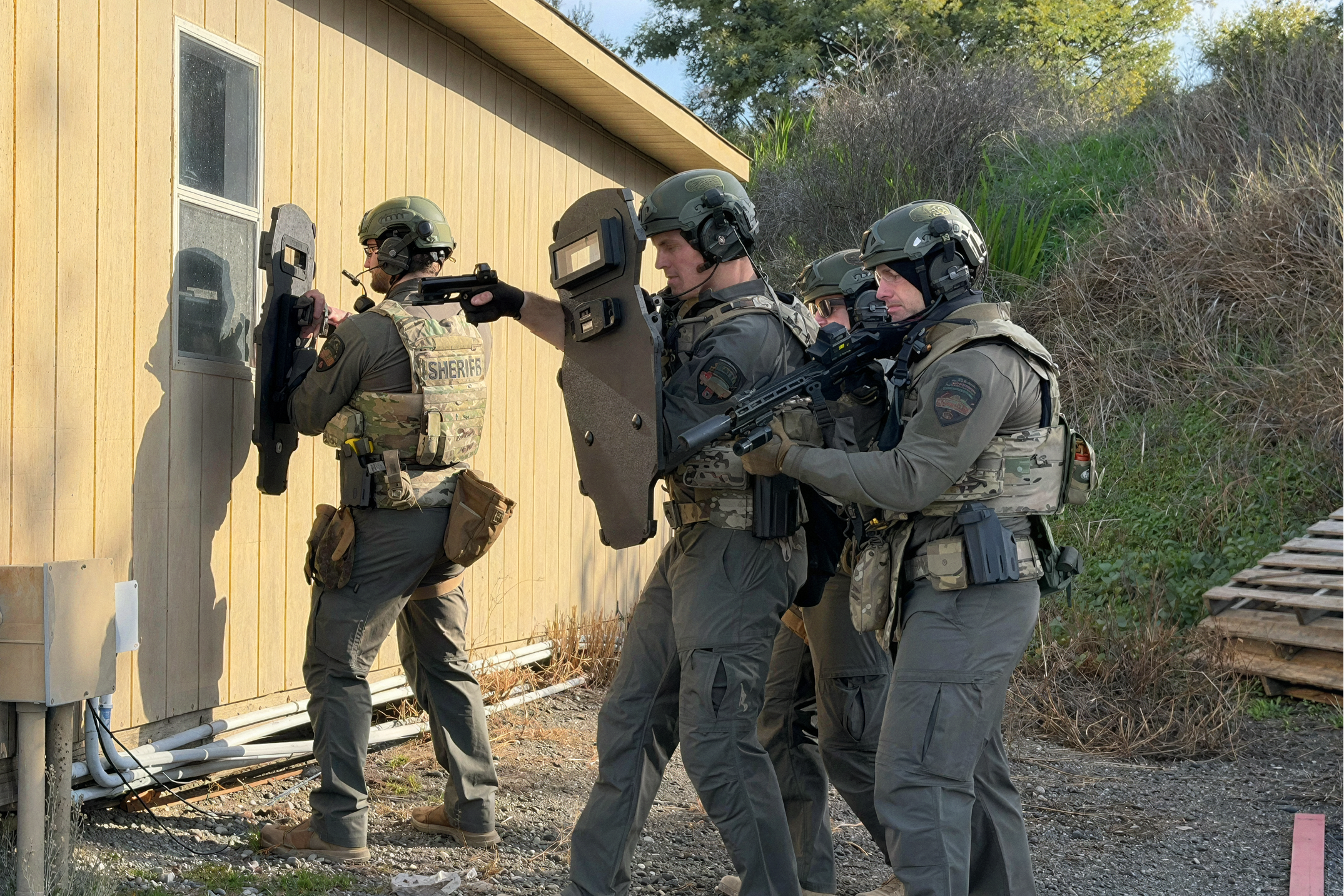 A tactical team of four officers in gear, armed and using shields, approaches a building's window, displaying caution and preparedness in a rural setting.