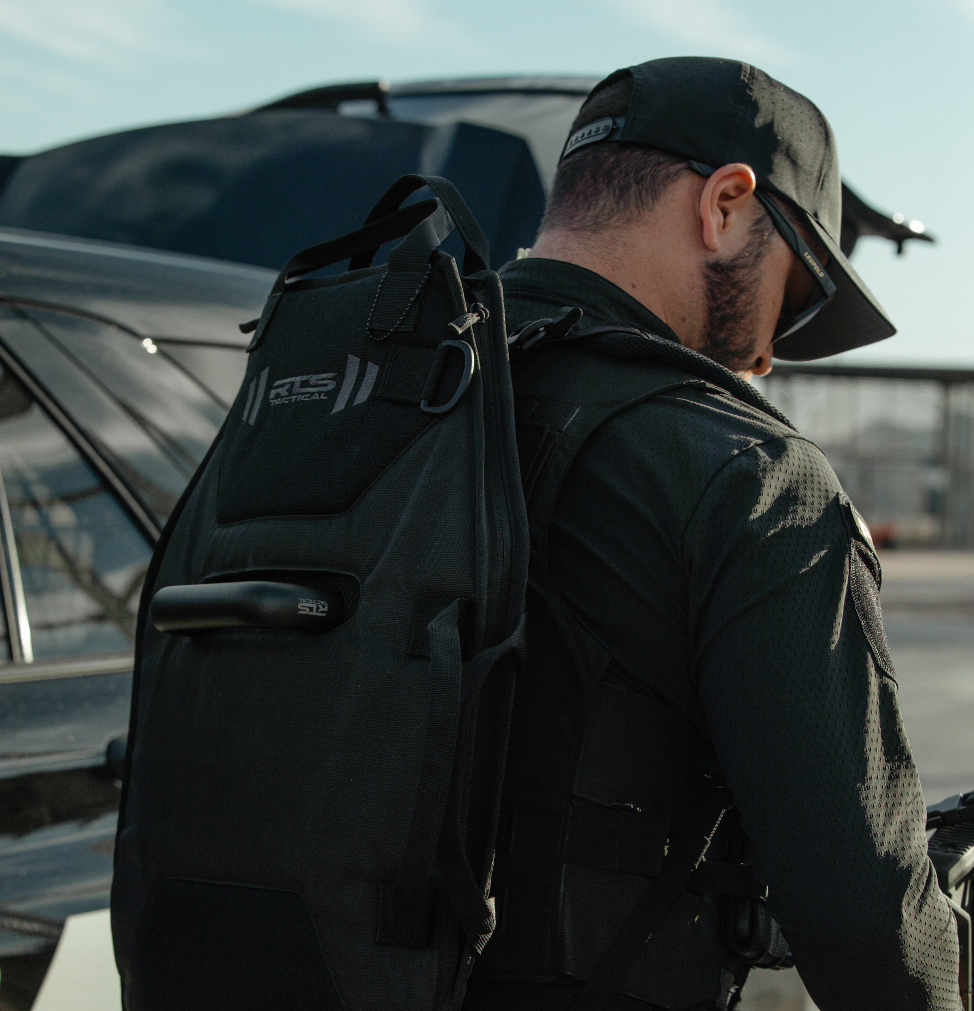 A man wearing a black tactical backpack and cap stands near a pickup truck, prepared for mission-ready operations.