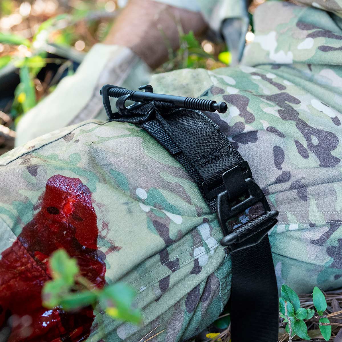 Close-up of a camouflage-clad leg with a black tourniquet applied above a bloodstained wound.