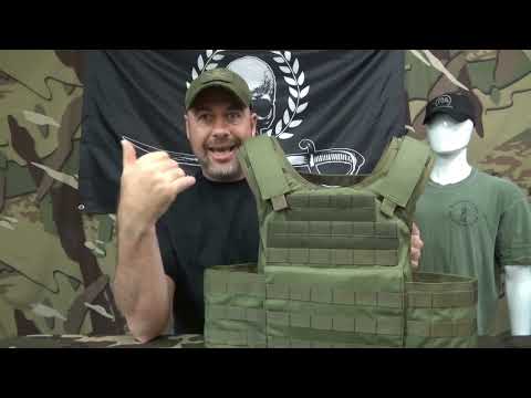 Man excitedly gestures with a thumbs-up next to a green tactical vest on a table. Camouflage background and mannequin with a cap and shirt nearby.