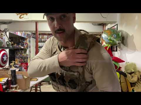 A man wearing a tactical vest stands in a cluttered room filled with colorful balloons, boxes, and various items.