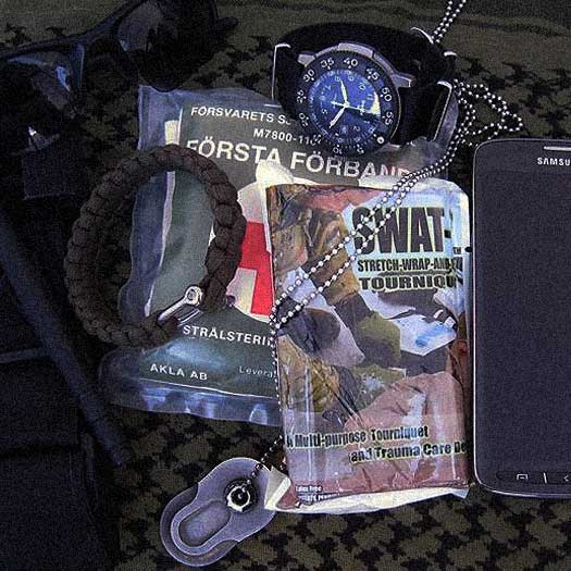 A collection of survival items on camouflage fabric, including sunglasses, a black watch, first aid kits, a smartphone, a paracord bracelet, and a dog tag.