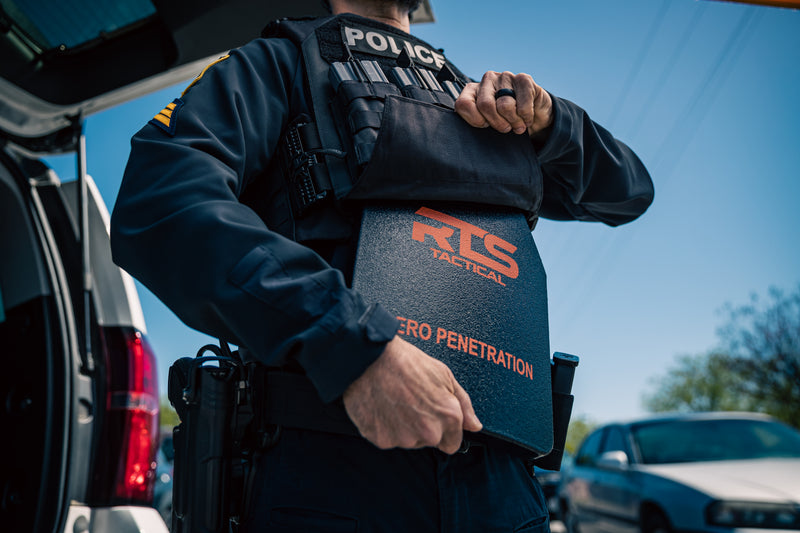 A police officer is securing a tactical ballistic shield with the label "ZERO PENETRATION" while standing near an open vehicle door.