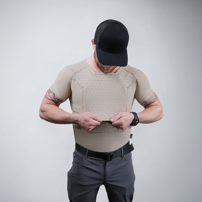 Man in tactical gear with a black cap and tattoo, adjusting a beige protective vest. Background is plain gray, conveying preparedness and focus.