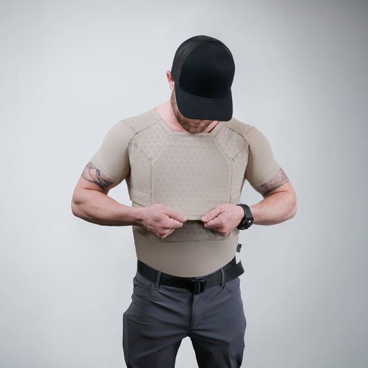 Man in tactical gear with a black cap and tattoo, adjusting a beige protective vest. Background is plain gray, conveying preparedness and focus.