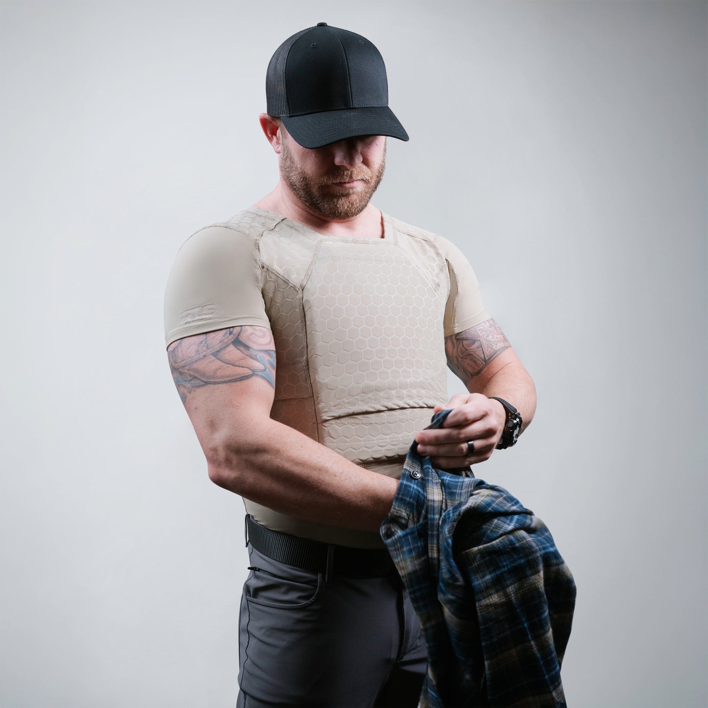 A man wearing a black cap and beige tactical vest holds a blue plaid shirt in his hands against a plain background.