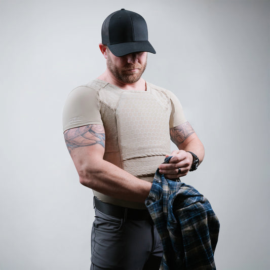 A man wearing a black cap and beige tactical vest holds a blue plaid shirt in his hands against a plain background.