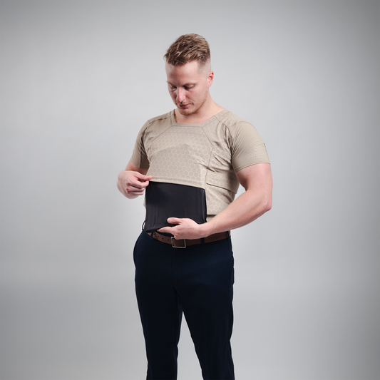 A man in a beige tactical shirt and dark pants adjusts a black protective vest on his torso. The background is plain gray, conveying focus and seriousness.