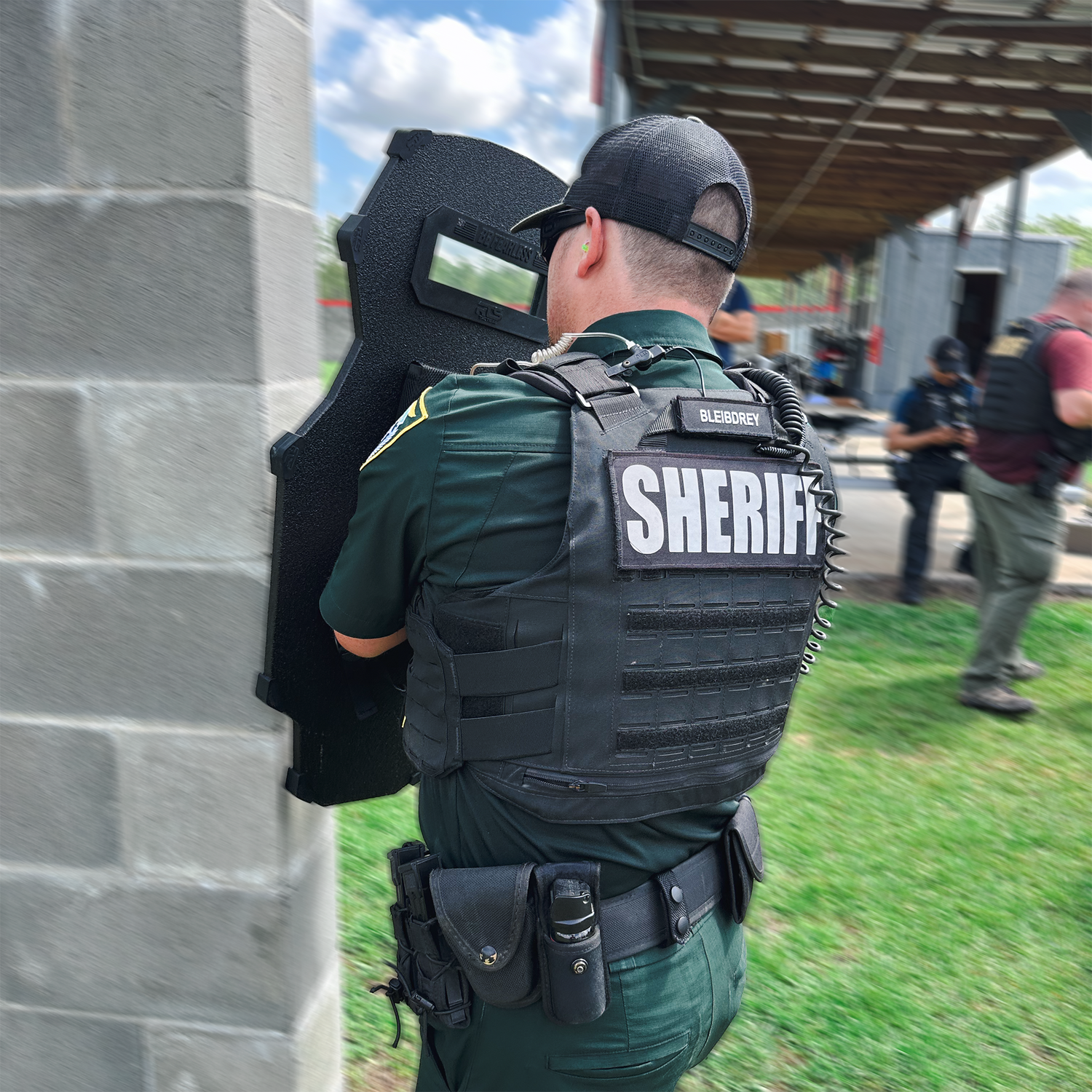 Sheriff officer in tactical gear holds a shield beside a concrete wall during a training session. Others in gear are seen in the background.