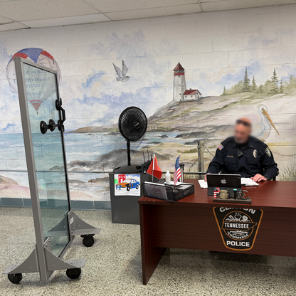 Police officer siting at a desk with a laptop, next to a bulletproof panel
