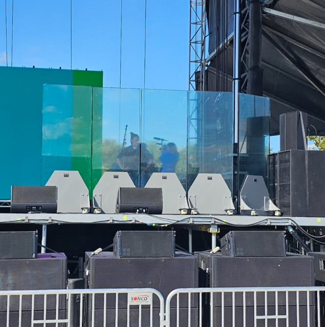 A stage setup with transparent sound barriers, amplifiers, and speakers. Musicians are faintly visible behind the barriers, under a clear blue sky.
