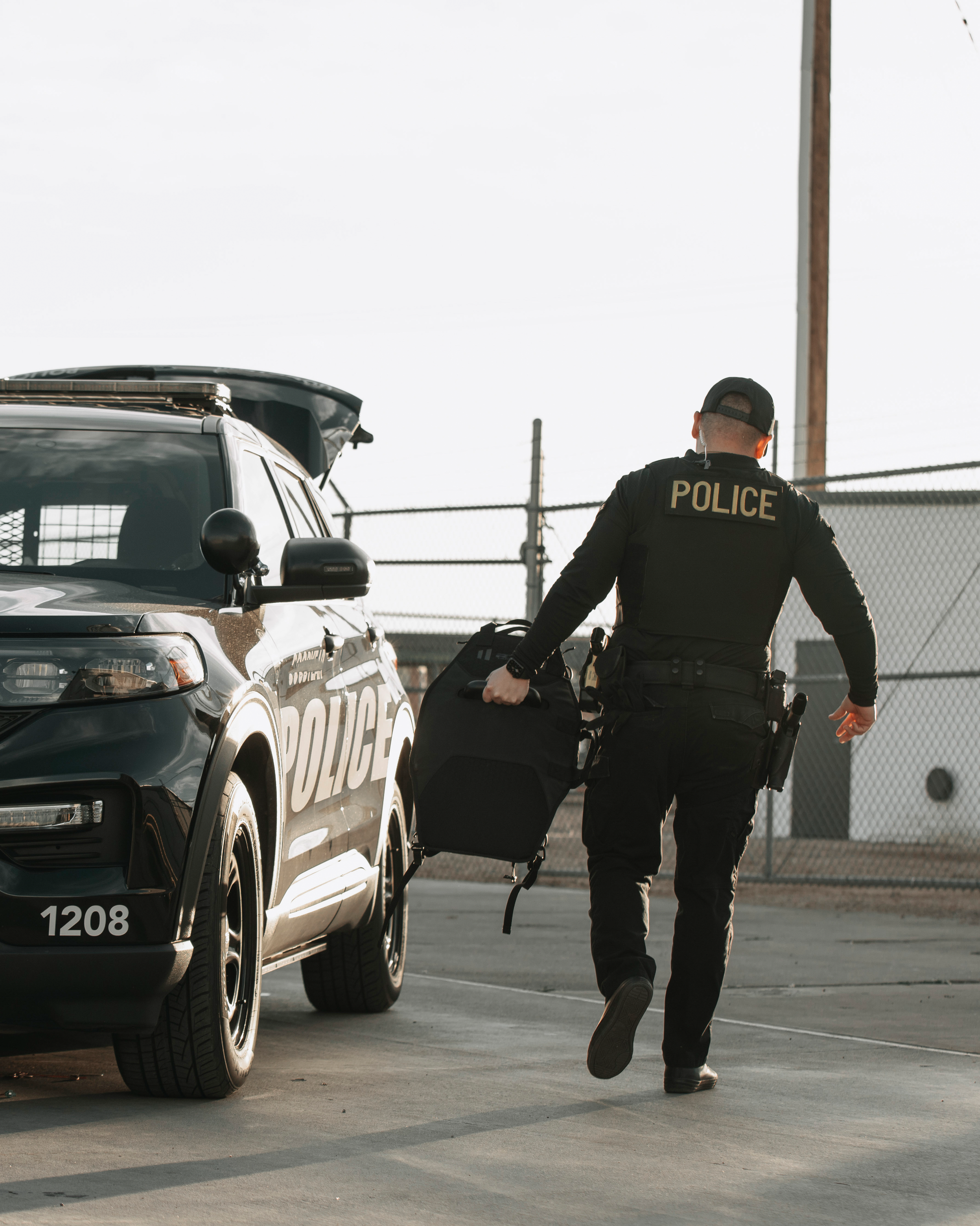 A police officer carries equipment from an SUV, with "POLICE" visible on the back of their uniform. The setting is an empty lot with a chain-link fence.