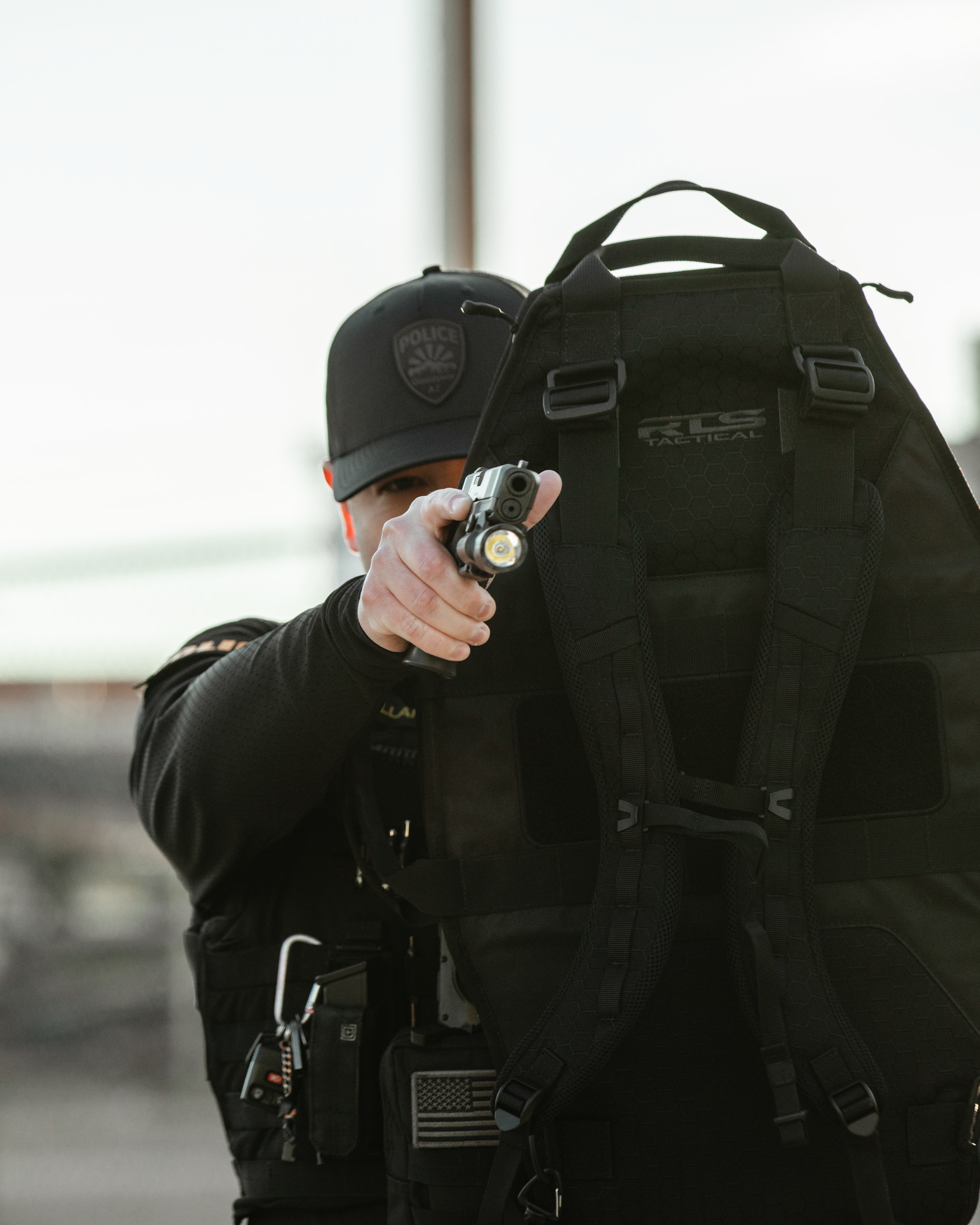 Police officer in tactical gear aims a flashlight-mounted firearm, partially obscured by a large black vest. The scene conveys readiness and intensity.