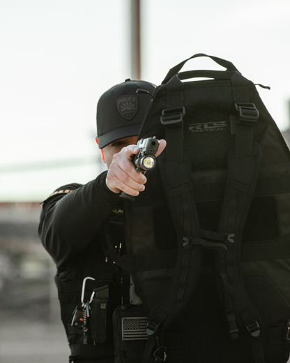 Police officer in tactical gear aims a flashlight-mounted firearm, partially obscured by a large black vest. The scene conveys readiness and intensity.