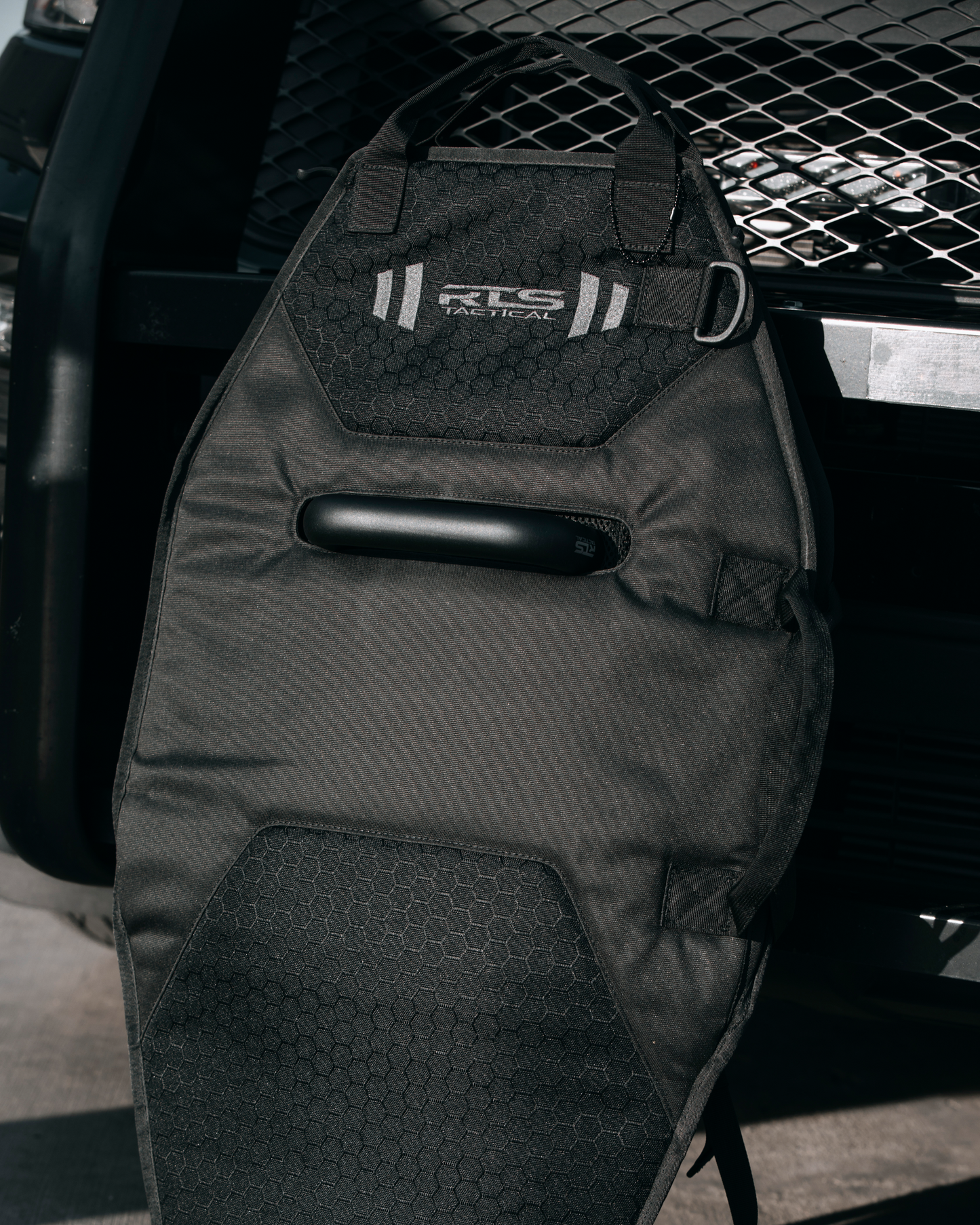 Black tactical bag for protective shield with hexagonal pattern and "RLS Tactical" logo, leaning against a metal grid surface, conveying strength and functionality.