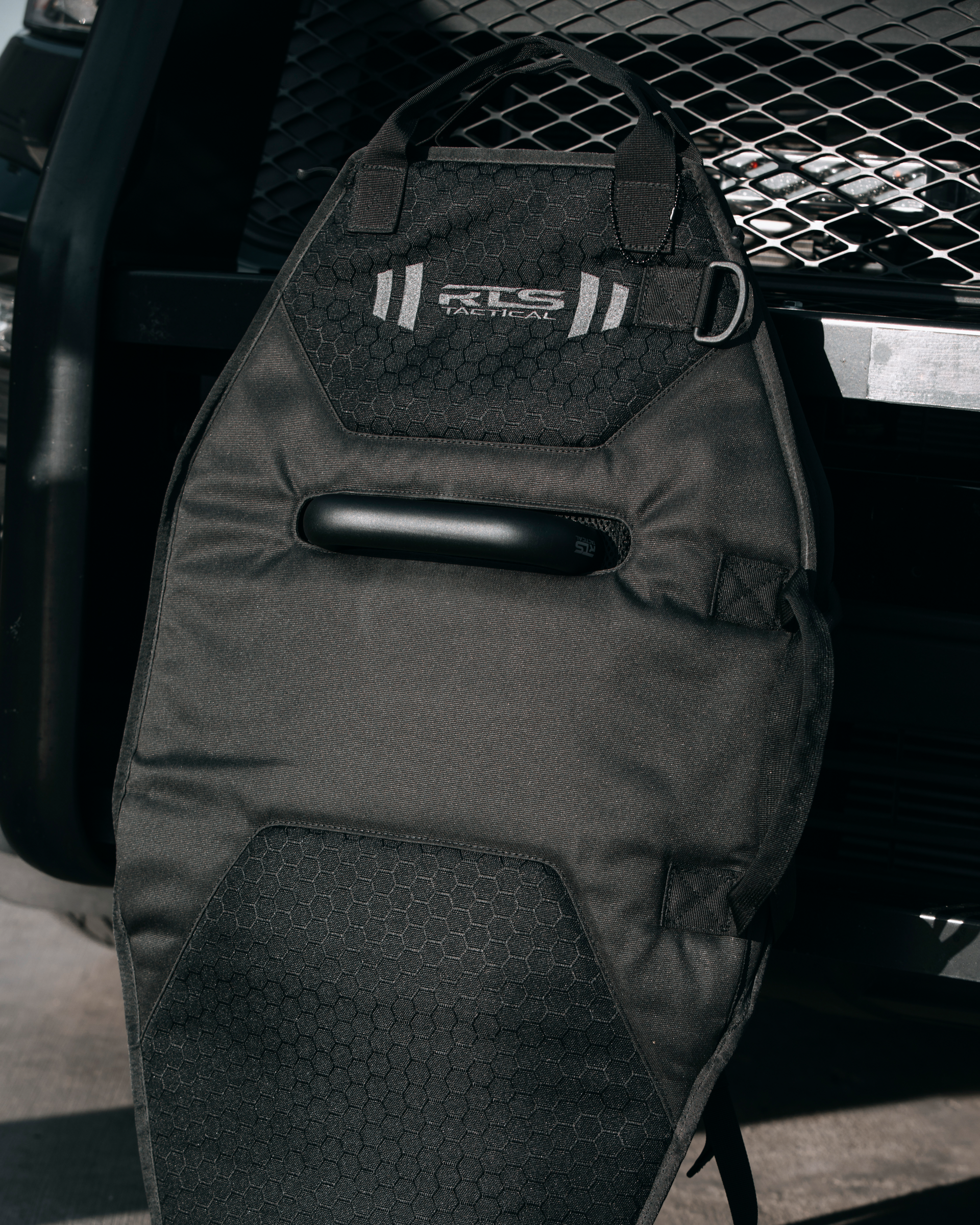 Black tactical bag for protective shield with hexagonal pattern and "RLS Tactical" logo, leaning against a metal grid surface, conveying strength and functionality.