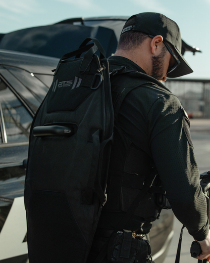 A person in tactical gear holds equipment, standing beside a vehicle. They wear a black cap and sunglasses, conveying readiness and focus.