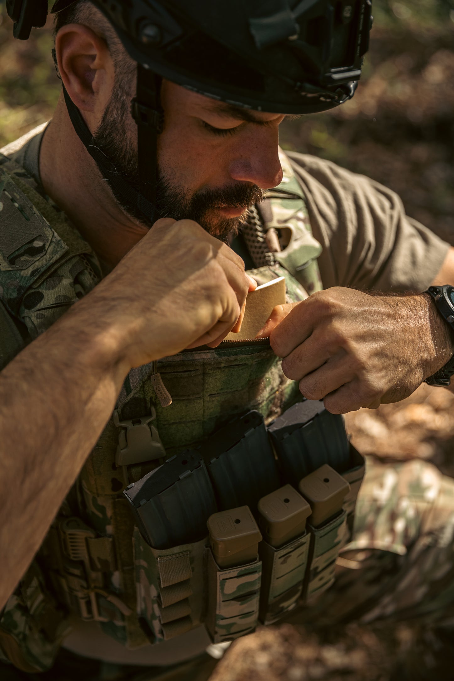 Man in tactical gear opens a pocket on his camouflage vest, revealing magazines. He wears a helmet, suggesting focus and readiness.