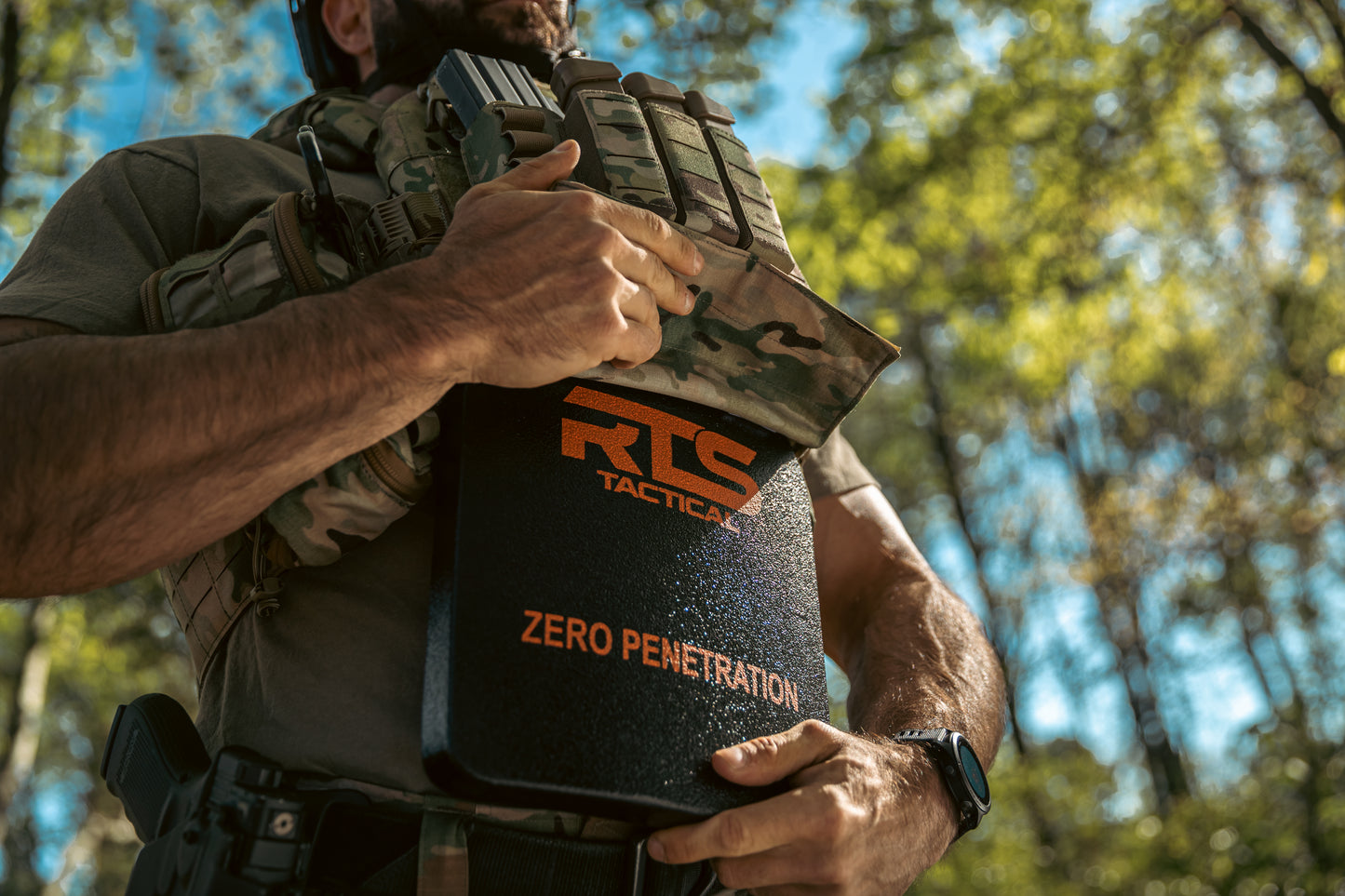 A man in tactical gear adjusts a vest in a sunlit forest. He's holding an armored plate labeled "RTS Tactical Zero Penetration."