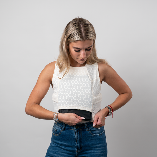 A woman in a white honeycomb-patterned compression shirt looks down while adjusting her waistband. She wears blue jeans and colorful bracelets, set against a plain gray background.