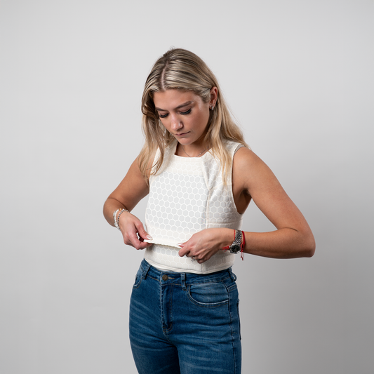 A woman in a sleeveless, textured compression shirt and blue jeans adjusts her shirt, focused. Her expression is neutral, standing against a plain gray background.