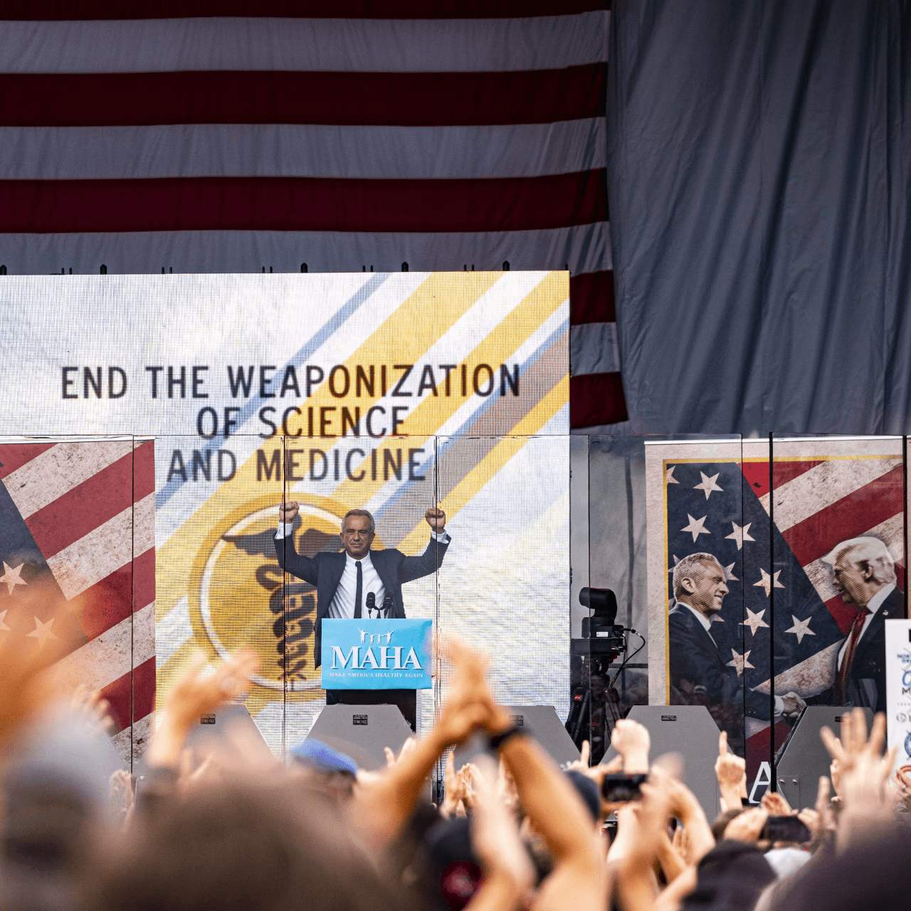 Robert F. Kennedy stands on stage at a rally, with a large U.S. flag backdrop. The screen reads, "End the weaponization of science and medicine."