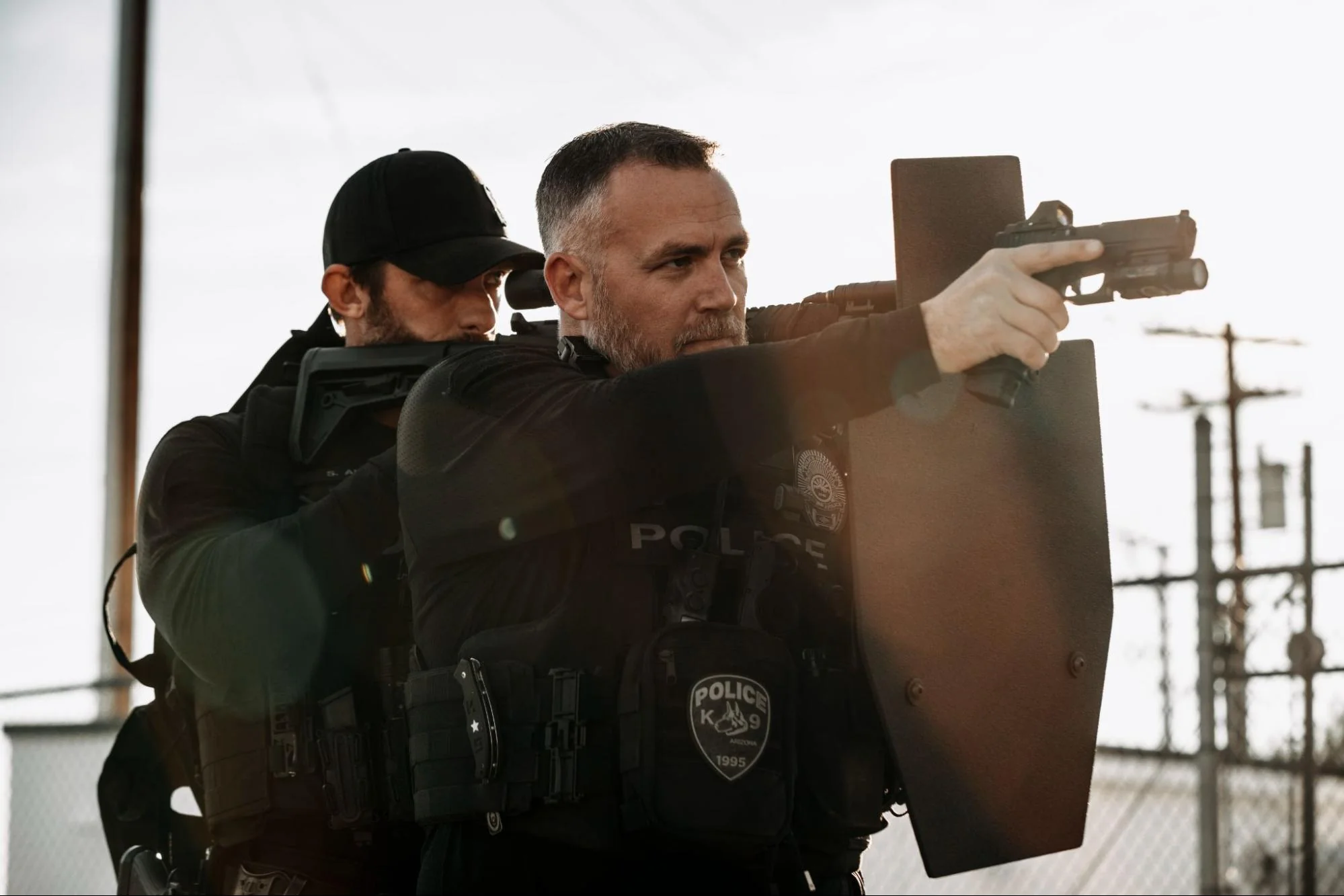 Two police officers in tactical gear are aiming firearms while taking cover behind a shield outdoors.