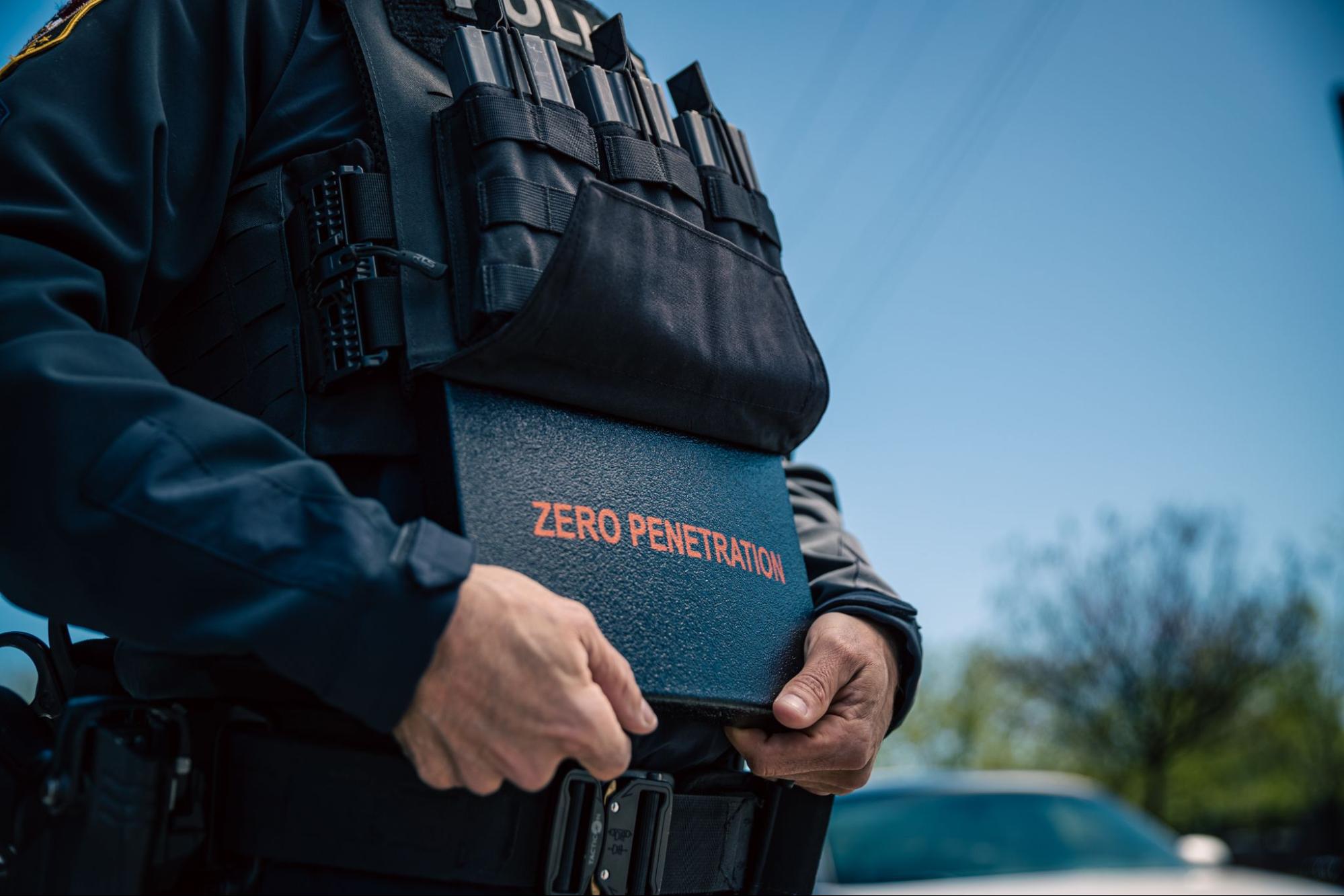 A tactical professional in uniform holds a blue ballistic plate carrier labeled "ZERO PENETRATION" while wearing duty gear and equipment.