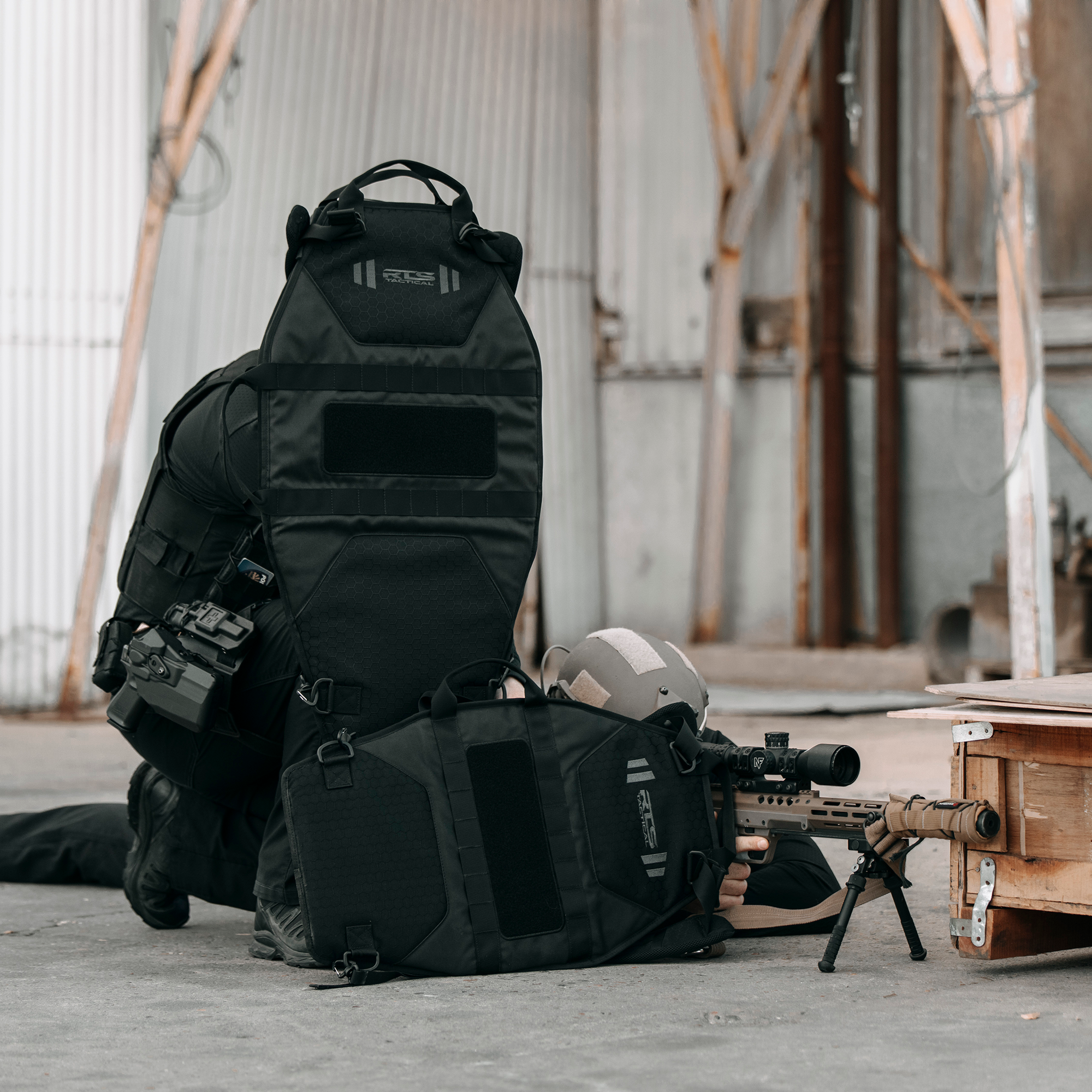 Tactical gear setup featuring black backpacks, a rifle with scope and bipod, ballistic helmet, and protective equipment arranged on concrete in an industrial warehouse setting.