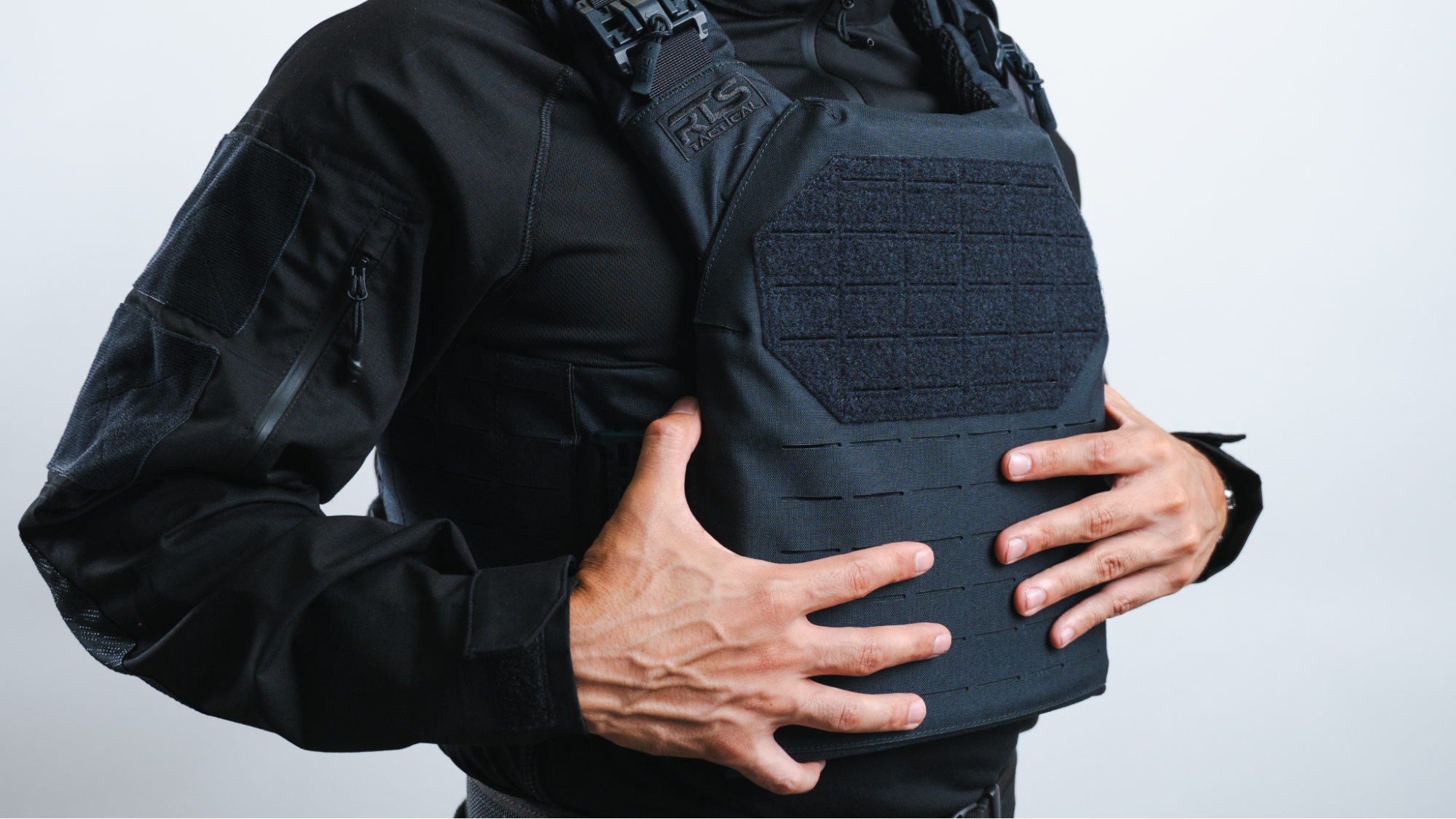 Person in tactical gear holding a black modular body armor plate carrier with MOLLE webbing.