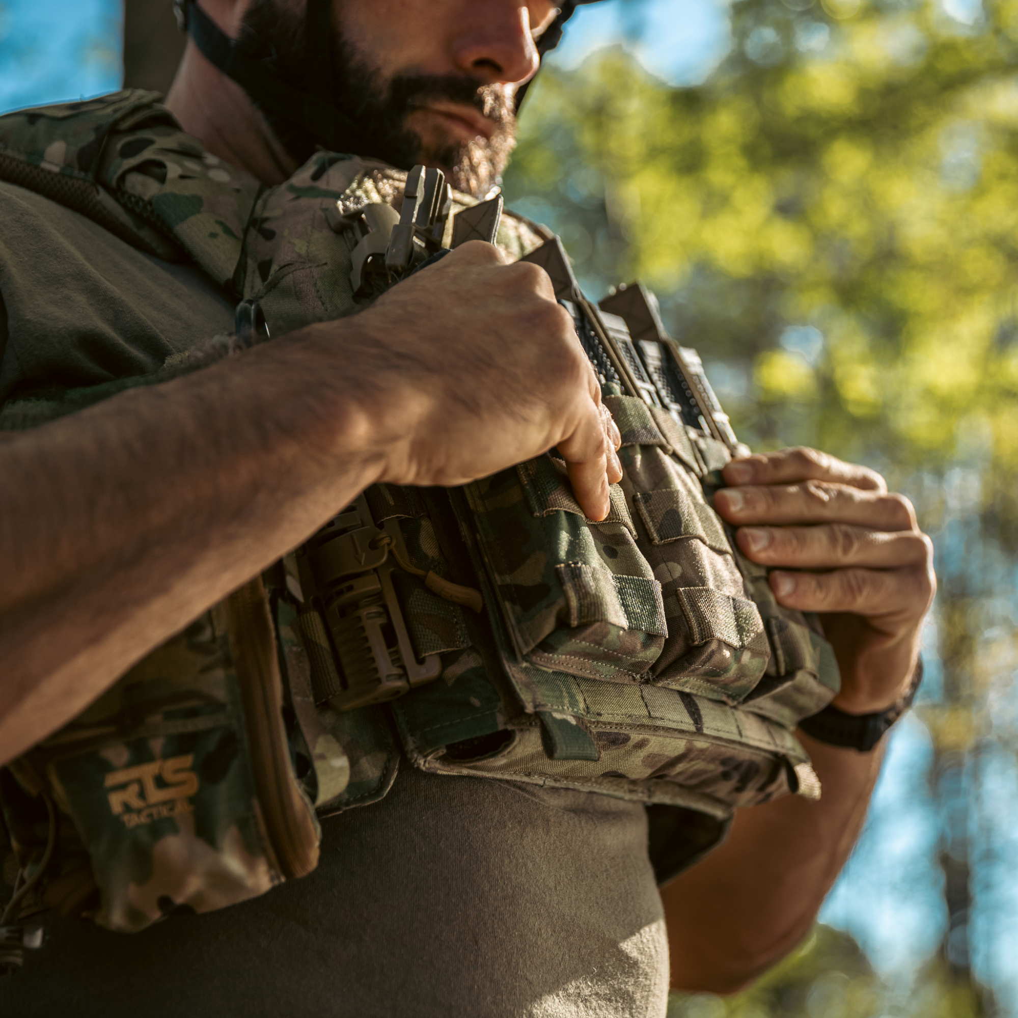 Bearded man in camouflage tactical gear adjusts his chest rig with multiple magazine pouches in an outdoor setting with trees in the background.