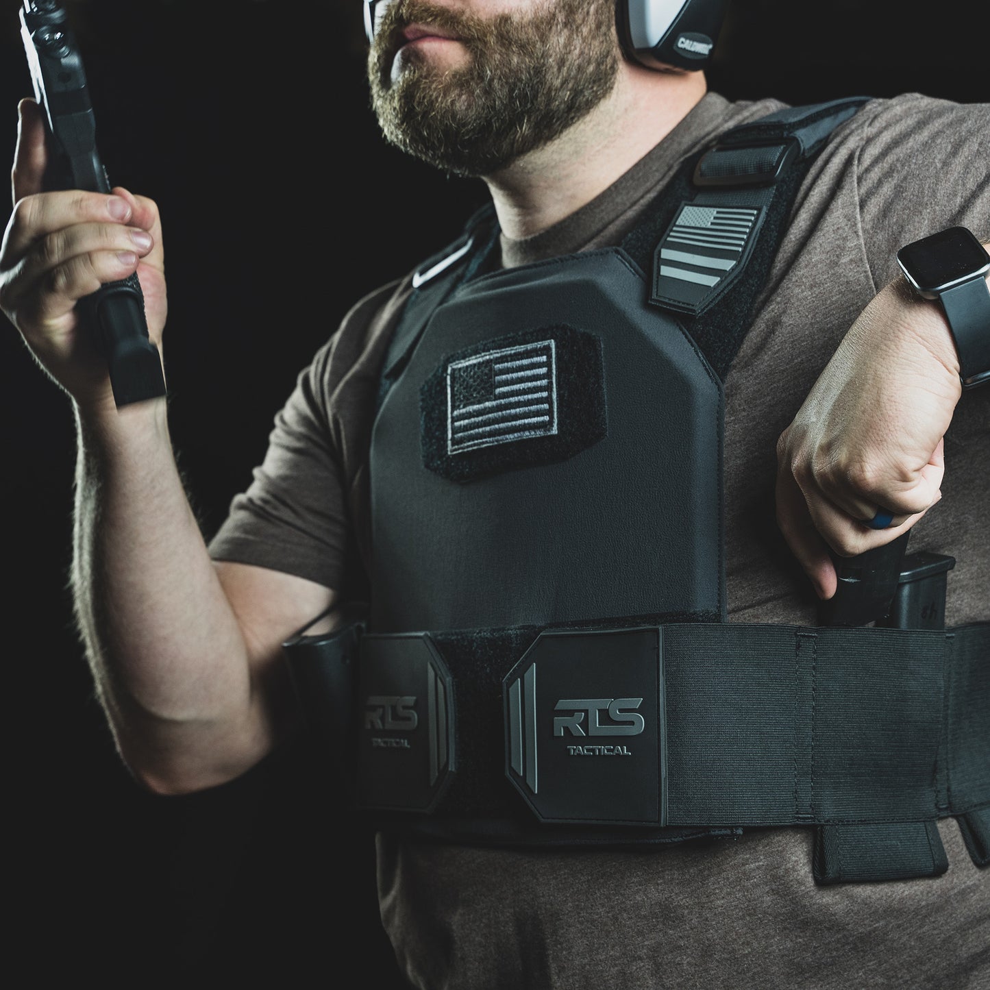 A man wearing a tactical vest holds a firearm in one hand while gripping a second weapon with the other.