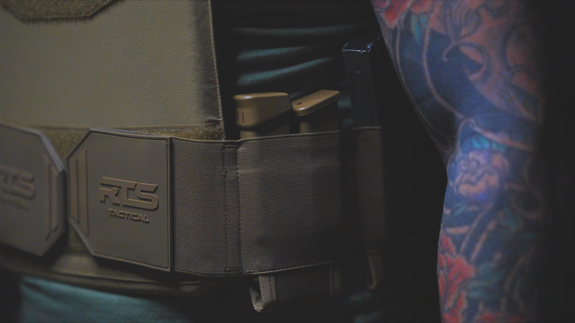 Close-up of a tactical vest with "RTS Tactical" logo, showcasing a magazine pouch holding two magazines. An arm with colorful tattoos is visible.