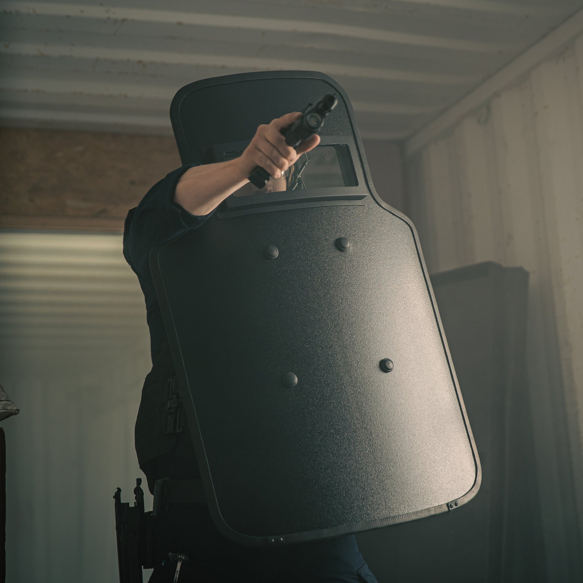 A person holding a handgun and aiming while standing behind a large ballistic shield in a dimly lit room.