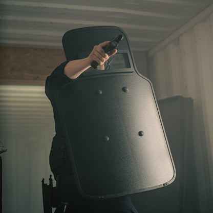 A person holding a handgun and aiming while standing behind a large ballistic shield in a dimly lit room.