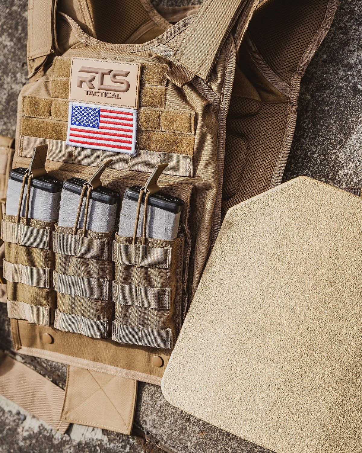 Tactical vest with pouches holding magazines, featuring a U.S. flag patch and "RTS Tactical" label. Beige armor plate beside it on a rough surface.