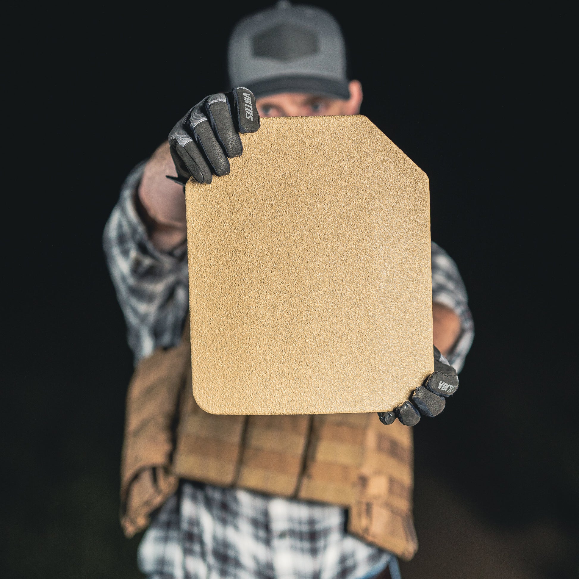 A person in tactical gear holds a rectangular bulletproof plate forward, focusing on the textured surface.
