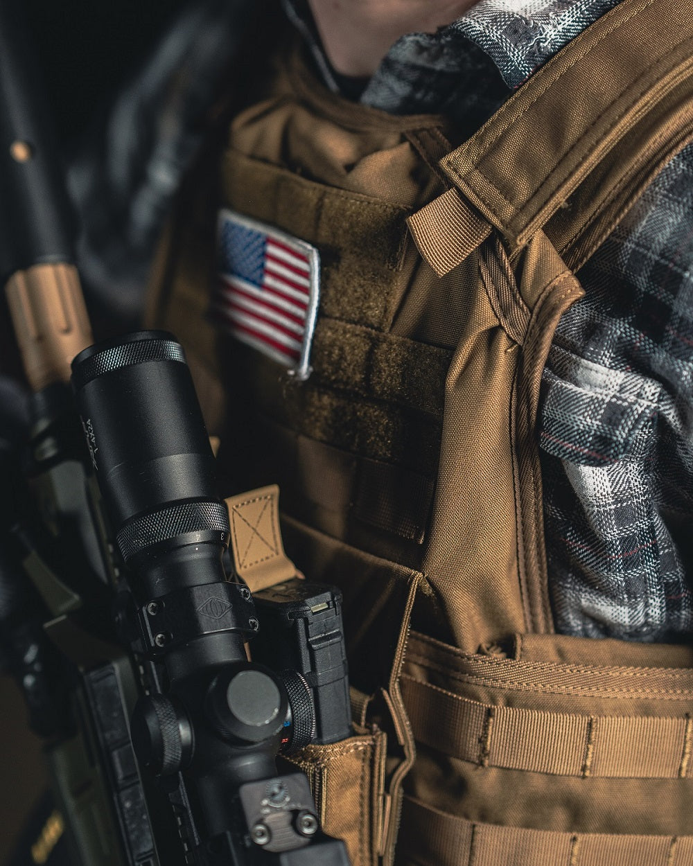 A person in a plaid shirt wears a tan tactical vest with a U.S. flag patch, holding a rifle with a scope.