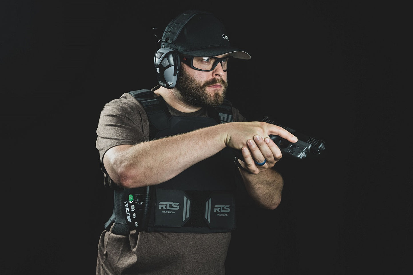 Man in tactical gear and headphones aiming a handgun, wearing protective glasses and a cap.