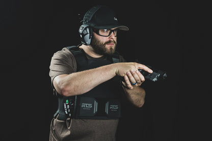 Man in tactical gear and headphones aiming a handgun, wearing protective glasses and a cap.