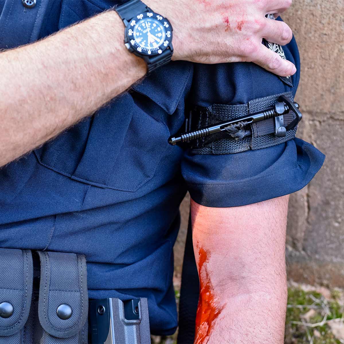 A police officer with a blue uniform applies a black tourniquet to their bloody arm, indicating an injury.