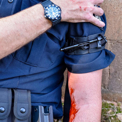 A police officer with a blue uniform applies a black tourniquet to their bloody arm, indicating an injury.